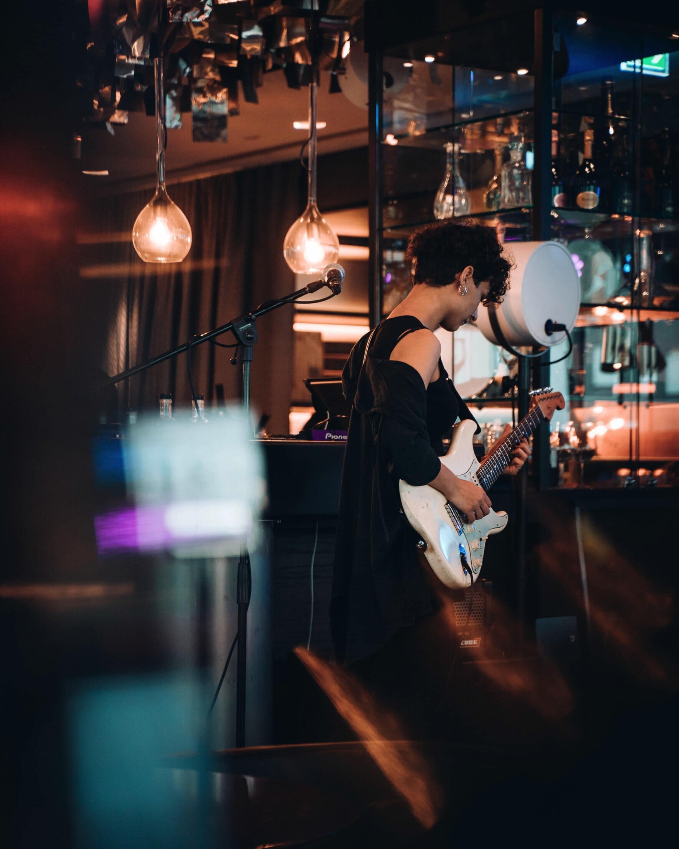 A person playing an electric guitar in a dimly lit room with hanging light fixtures and a bar in the background.