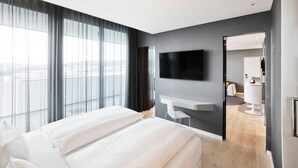 A modern hotel room with a large bed, a flat-screen TV mounted on the wall, a small table, and a view of the city through large windows. - Jaz in the City Stuttgart (Stuttgart)
