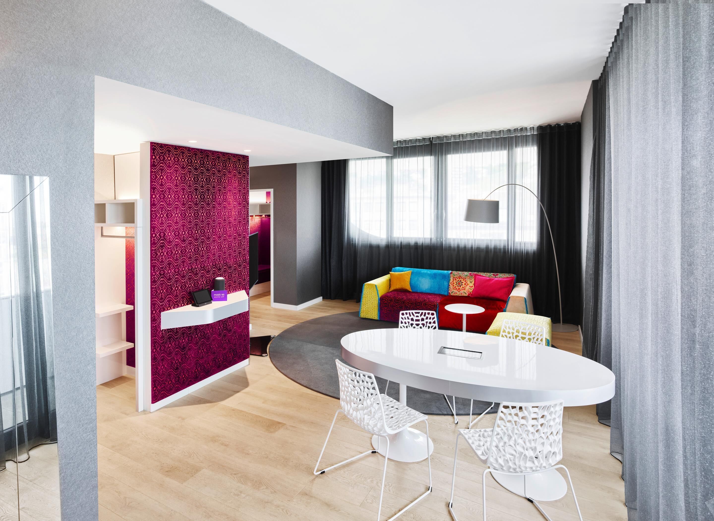 A modern hotel room with a round dining table, white chairs, a colorful sofa, a desk with a computer, and a large window with curtains.