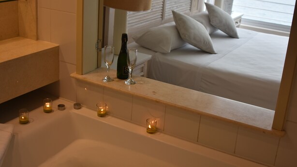 A bathroom with a bathtub, a mirror, and a bed with white bedding and pillows. There are candles on the bathtub and a bottle of champagne on the bed. - Hippocampus Resort & Club (Concon)