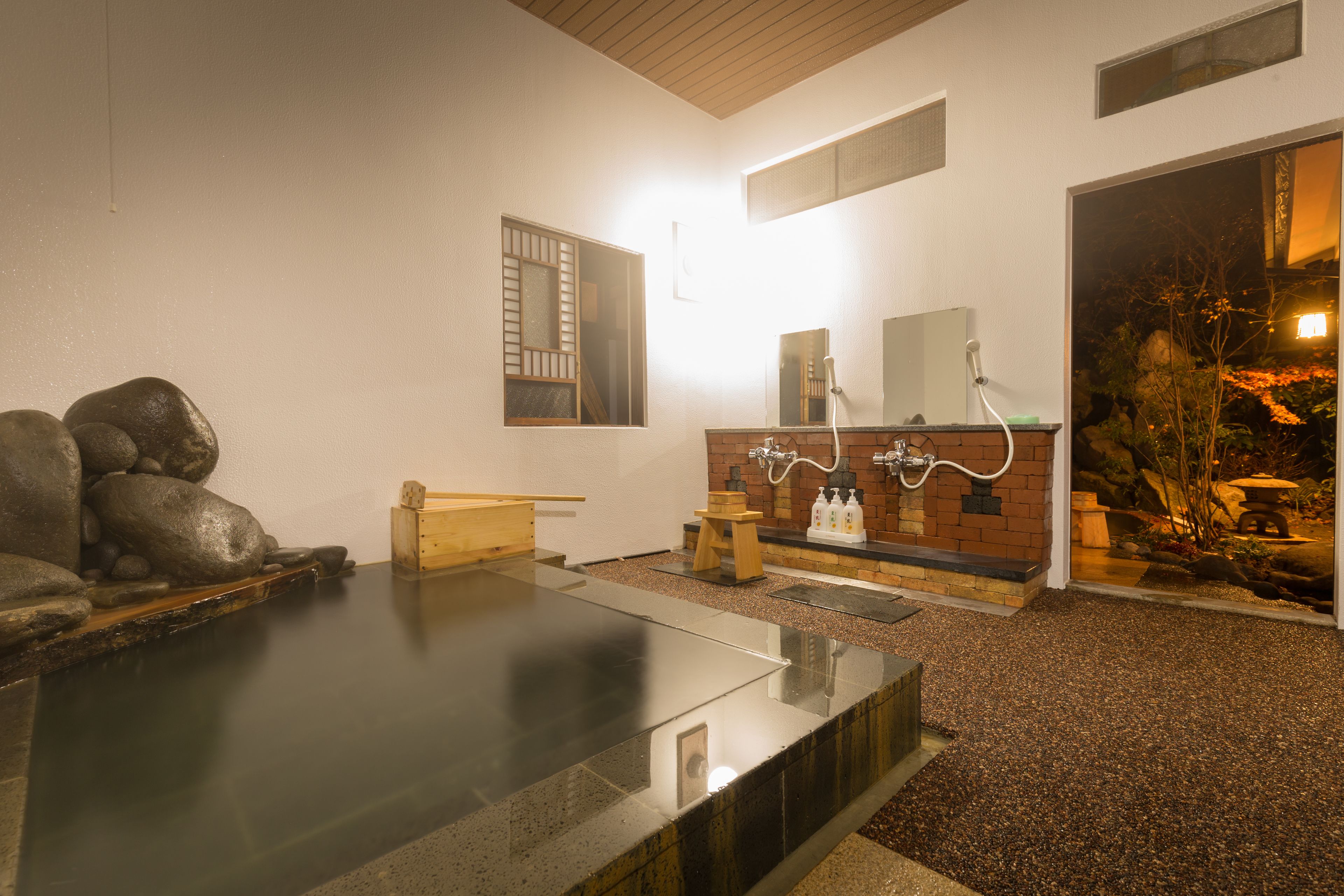 A traditional Japanese onsen with a stone hot spring, a wooden bench, a brick counter with a mirror, and a small garden area visible through an open doorway.