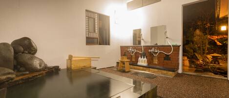 A traditional Japanese onsen with a stone hot spring, a wooden bench, a brick counter with a mirror, and a small garden area visible through an open doorway.