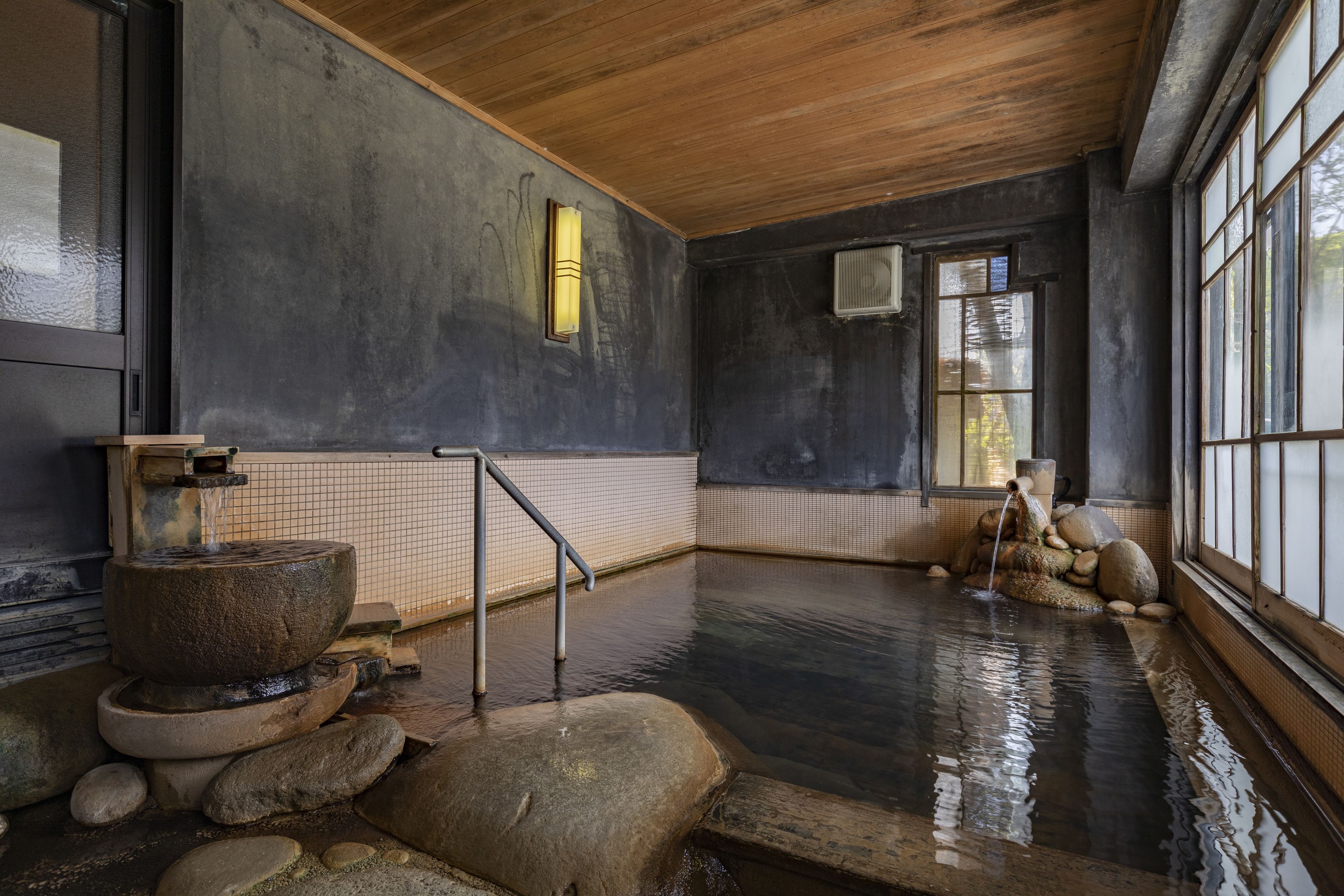 Un onsen japonais traditionnel, avec une source d’eau chaude alimentée par des pierres, des poutres en bois et des murs carrelés.