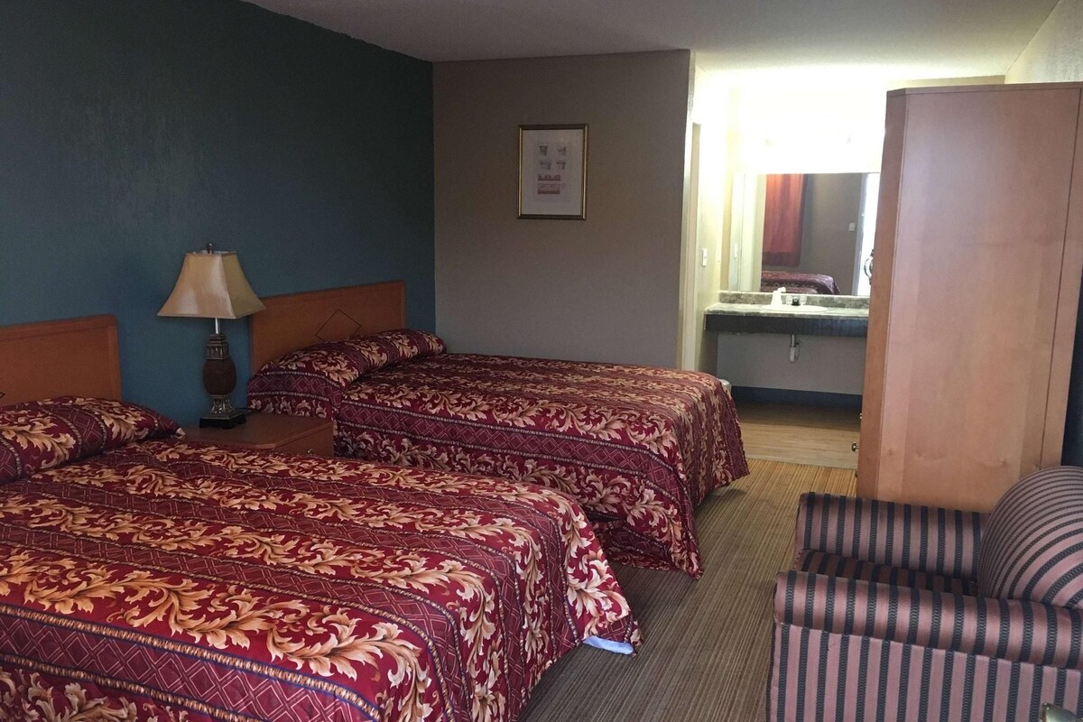 A hotel room with two beds, a chair, a lamp, a mirror, and a bathroom visible through an open door.