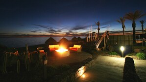 Een terras aan het strand met zitplaatsen en een vuurkorf, uitkijkend over de oceaan bij zonsondergang.