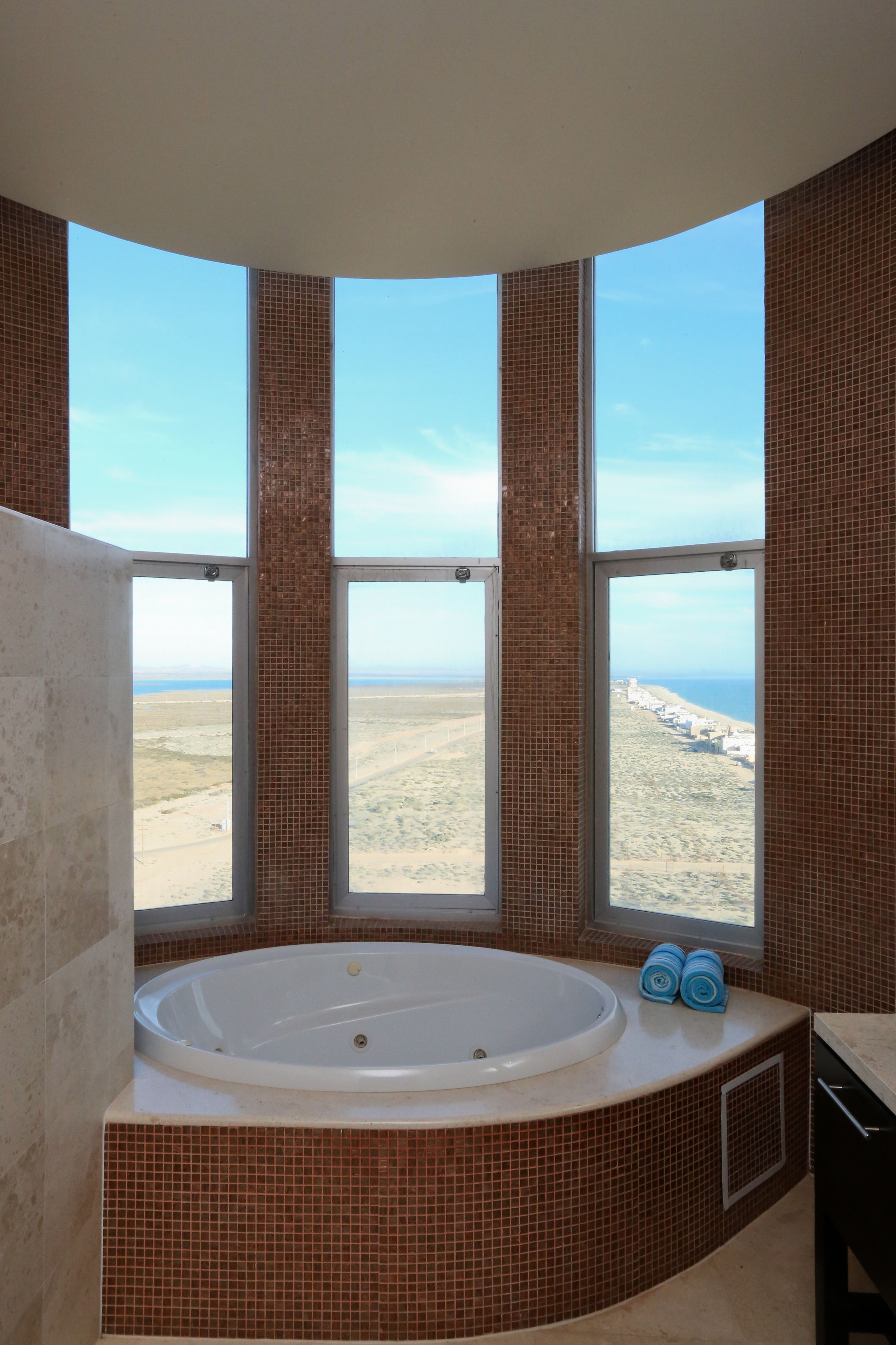 A bathroom with a large bathtub, a window with a view of a beach, and a tiled wall.