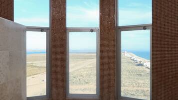 A bathroom with a large bathtub, a window with a view of a beach, and a tiled wall.