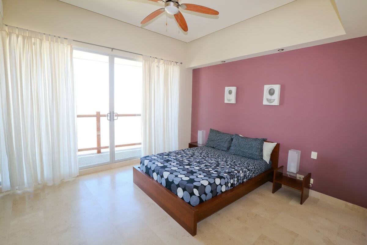 Un dormitorio con cama, cortinas, ventilador de techo, mesita de noche y lámpara de pared.