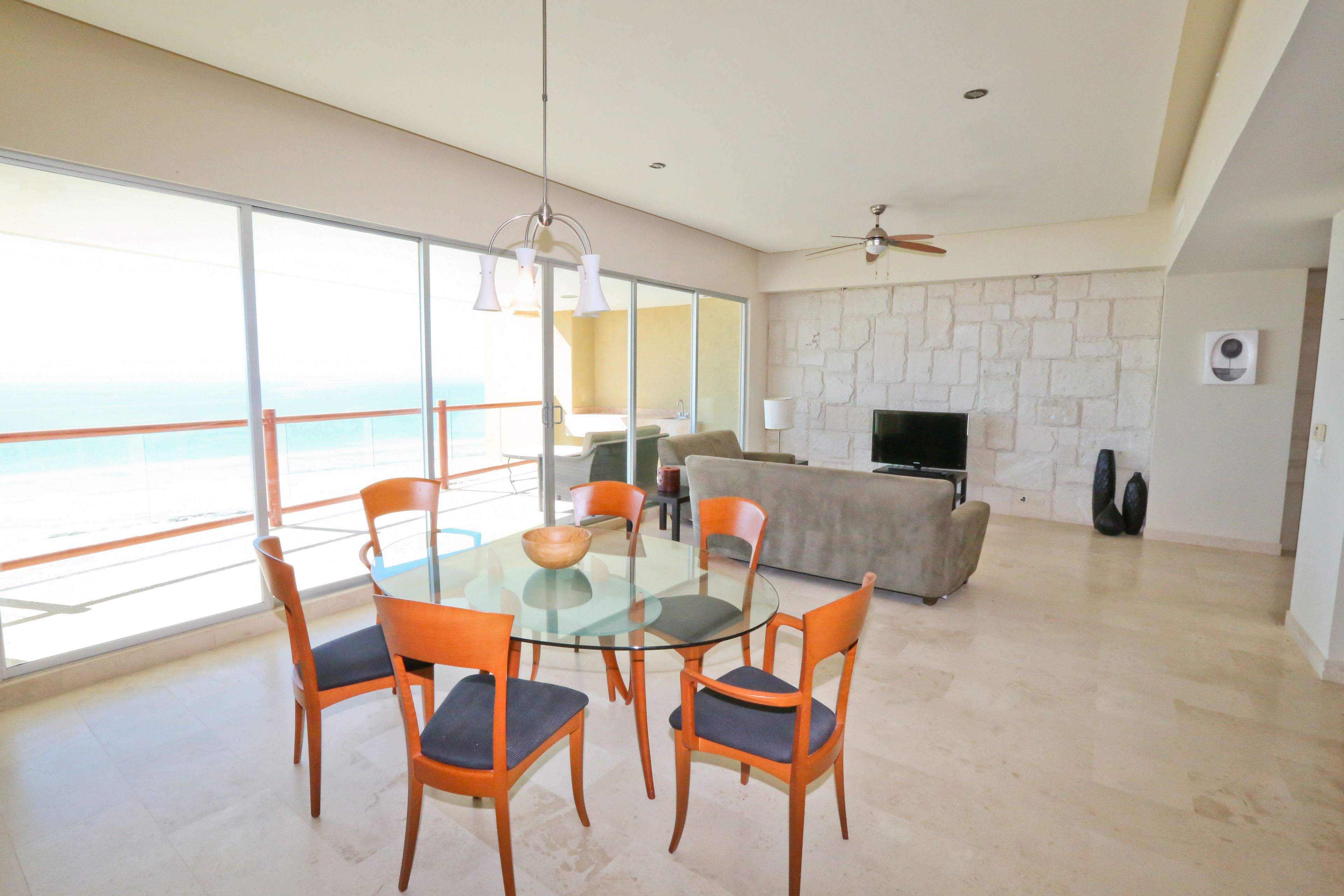 A modern living room with a glass dining table, orange chairs, a beige sofa, and a TV.
