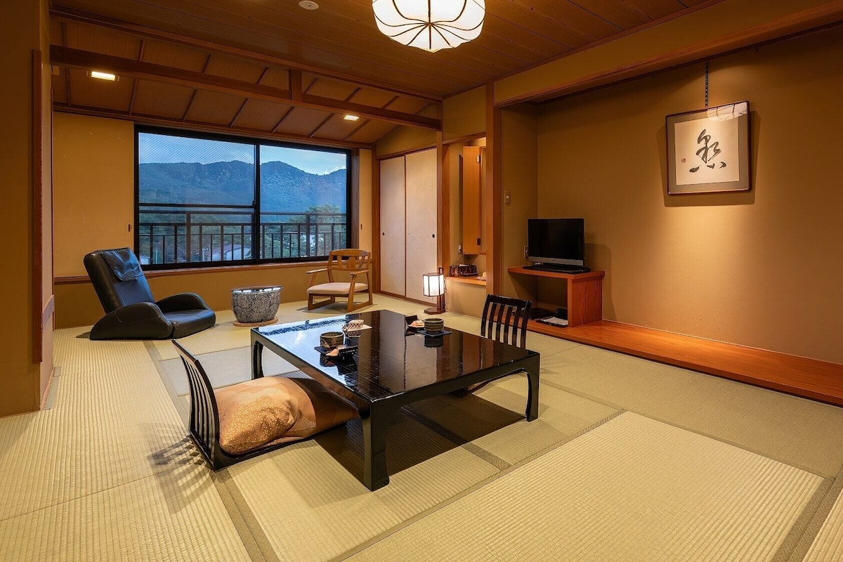 Une pièce de style japonais traditionnel, avec un sol recouvert de tatamis, une table basse, un canapé, un téléviseur et une vue sur les montagnes par la fenêtre.