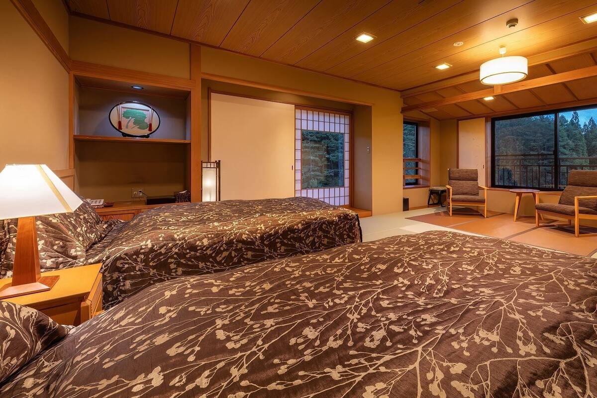 Une chambre de style japonais traditionnel, avec un sol recouvert de tatamis, un grand lit avec une literie à motifs, un bureau avec une lampe et une vue sur une forêt, visible à travers de grandes fenêtres.
