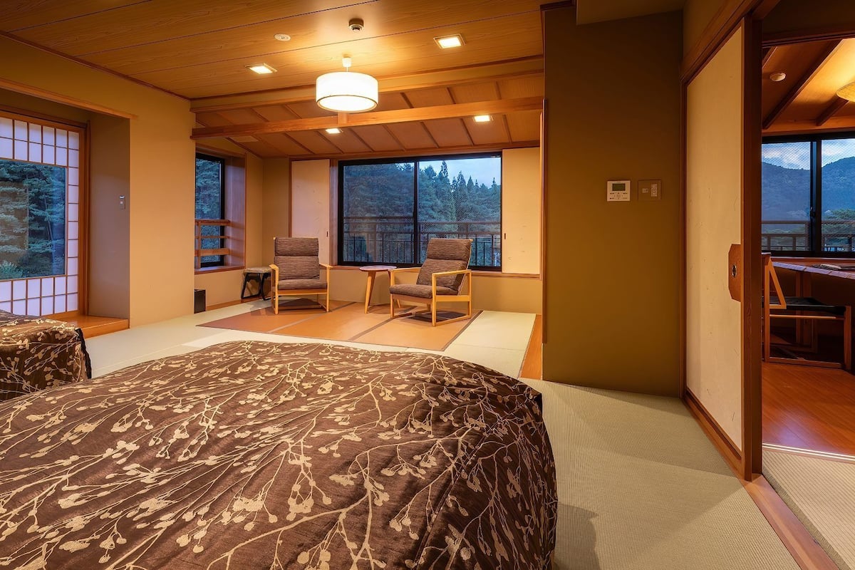 Une pièce de style japonais traditionnel, avec un sol recouvert de tatamis, un lit agrémenté d’une couvre-lit à motifs, un bureau et une vue sur une forêt, visible à travers de grandes fenêtres.
