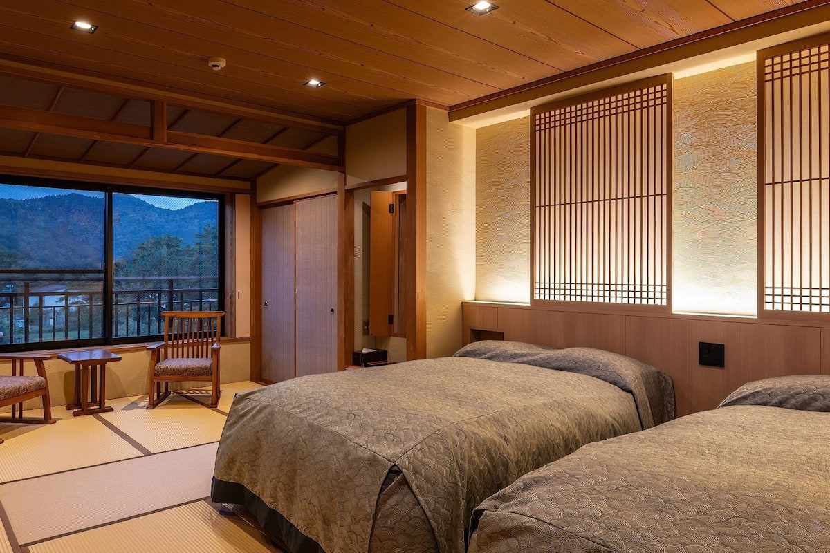 Une pièce de style japonais traditionnel, avec un sol recouvert de tatamis, un lit bas, un bureau en bois avec des chaises et une grande fenêtre donnant sur une vue sur la montagne.