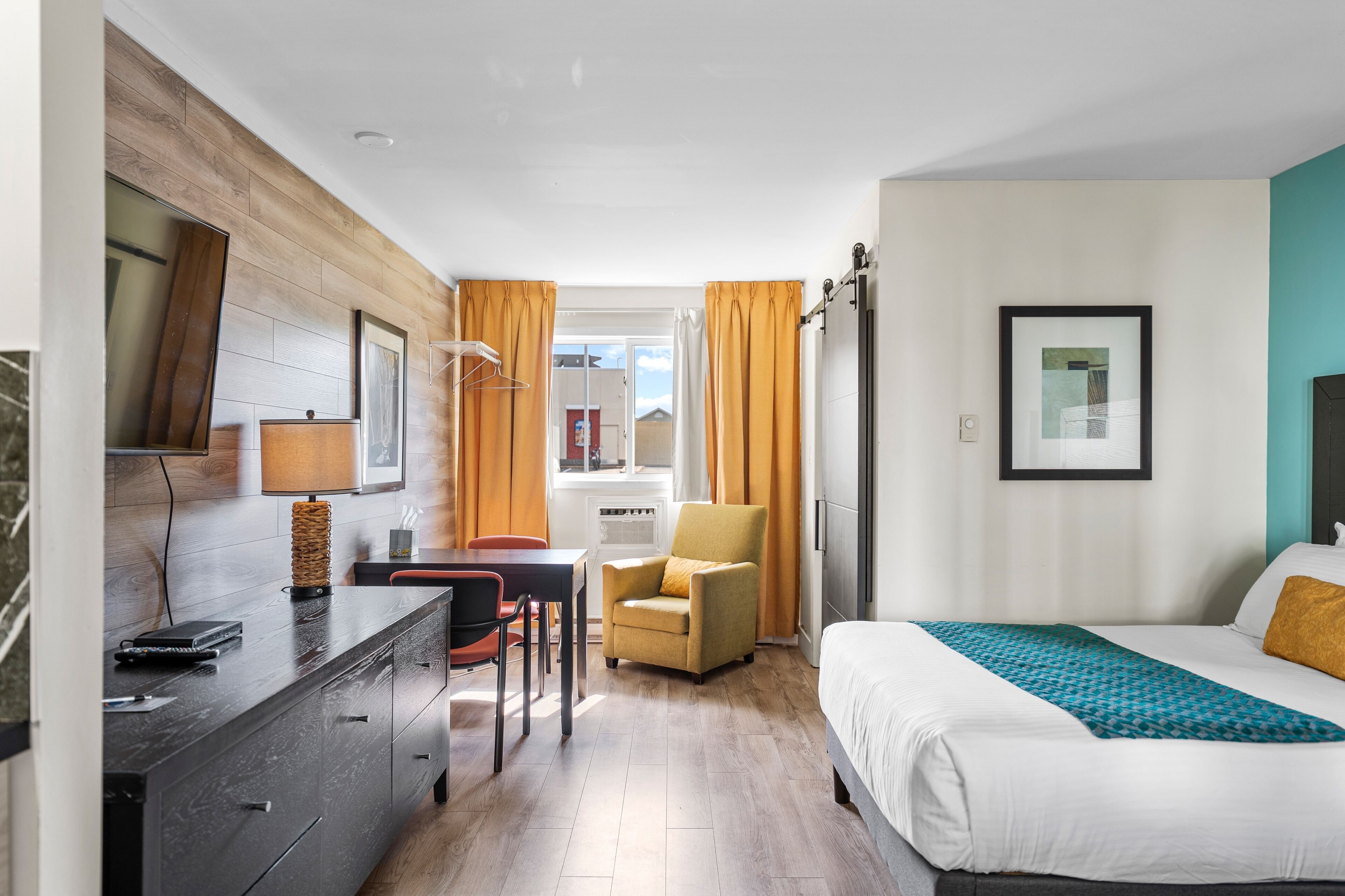 A hotel room with a bed, a desk with a lamp, a chair, and a wardrobe.