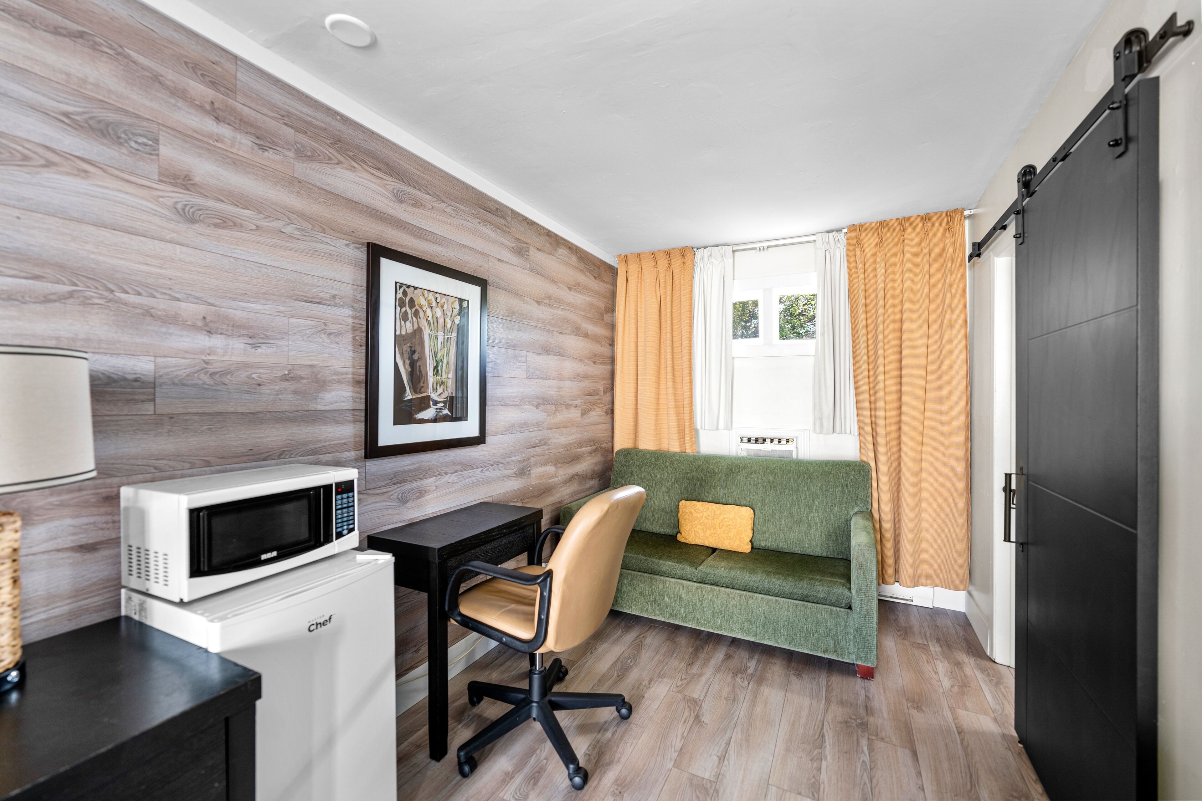 Habitación compacta con pared de madera, un sofá verde, un escritorio blanco con microondas y una puerta corredera.