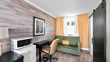 Habitación compacta con pared de madera, un sofá verde, un escritorio blanco con microondas y una puerta corredera.