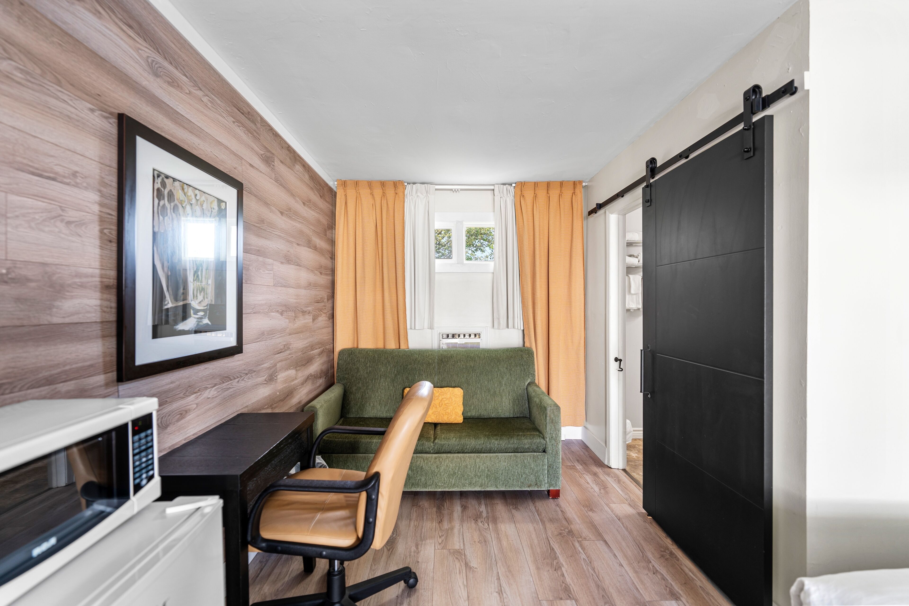 Una habitación compacta con un sofá verde, piso de madera, microondas blanco, escritorio negro y un cuadro en la pared.