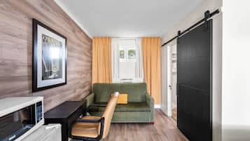 Una habitación compacta con un sofá verde, piso de madera, microondas blanco, escritorio negro y un cuadro en la pared.