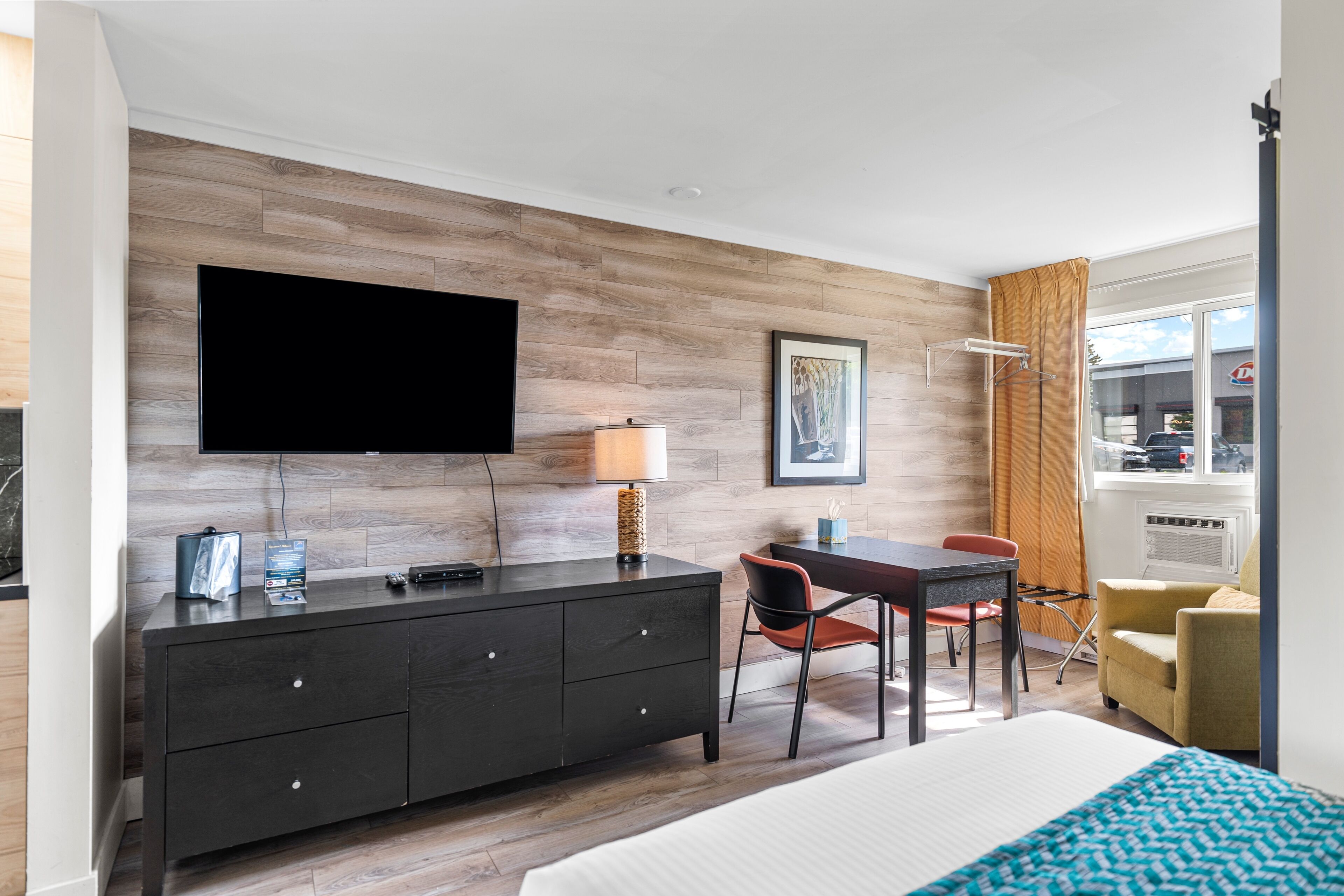 A modern hotel room with a flat-screen TV, a wooden accent wall, a desk with a chair, a bed with a blue and white patterned blanket, and a window with curtains.