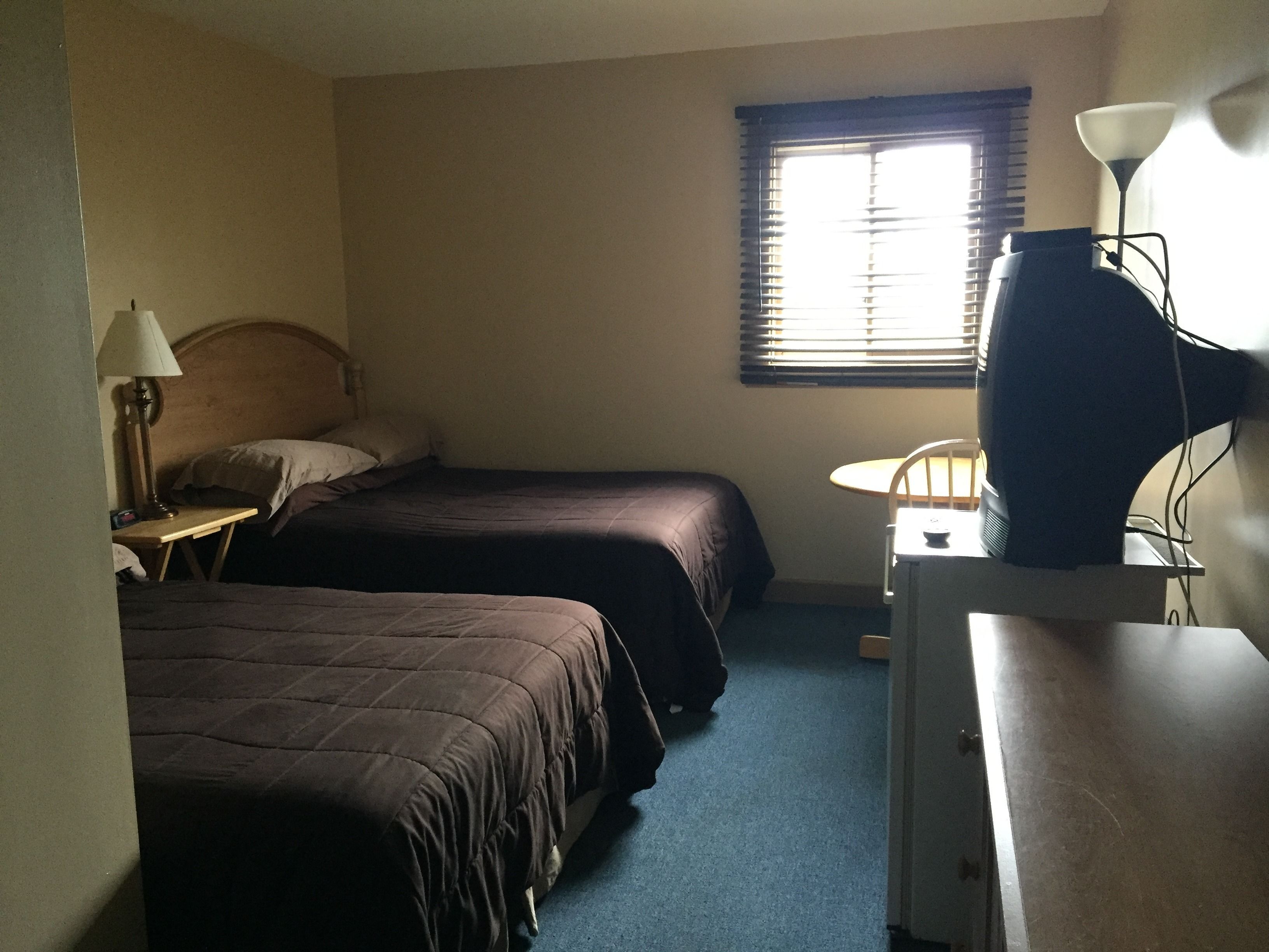A small, dimly lit room with two beds, a desk, and a window.