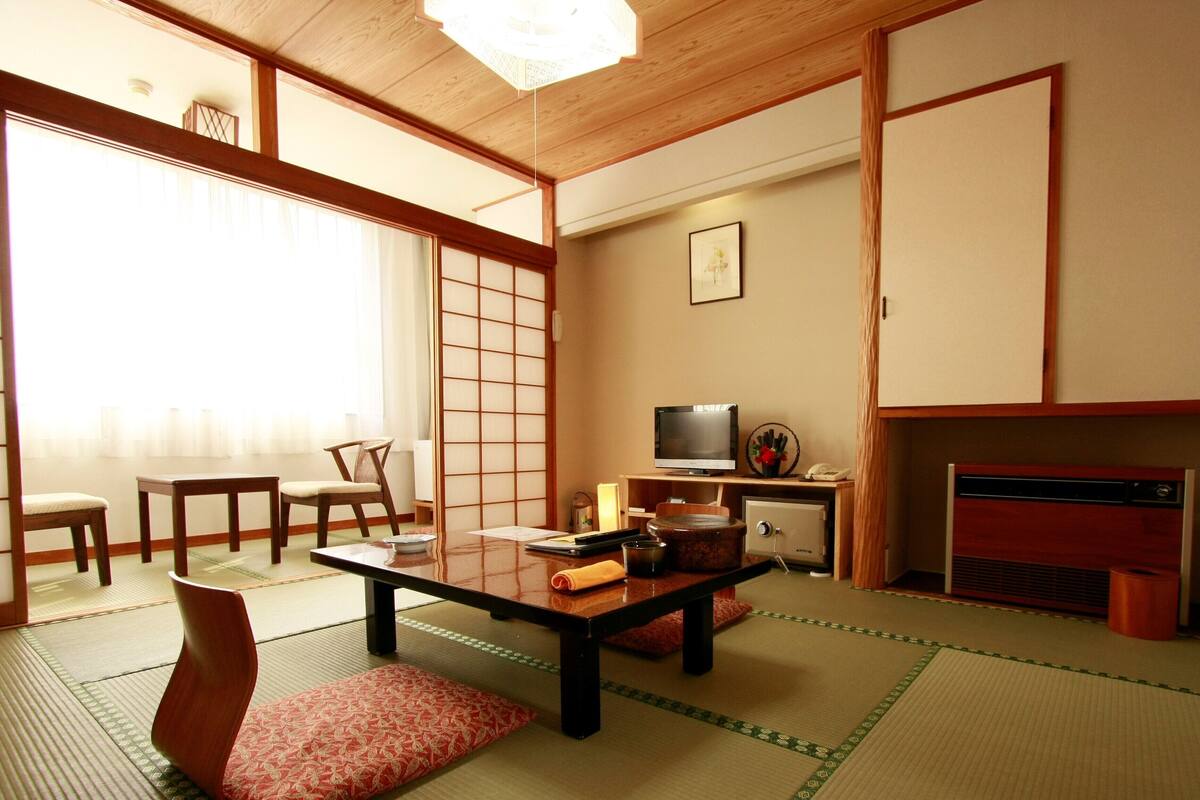 A traditional Japanese room with tatami flooring, a low table, chairs, a television, and a fireplace.