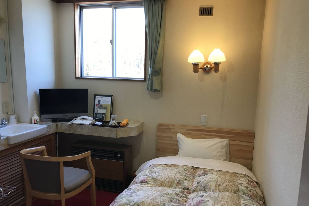 A hotel room with a bed, a desk with a computer, a chair, a window with curtains, and a wall-mounted light fixture.