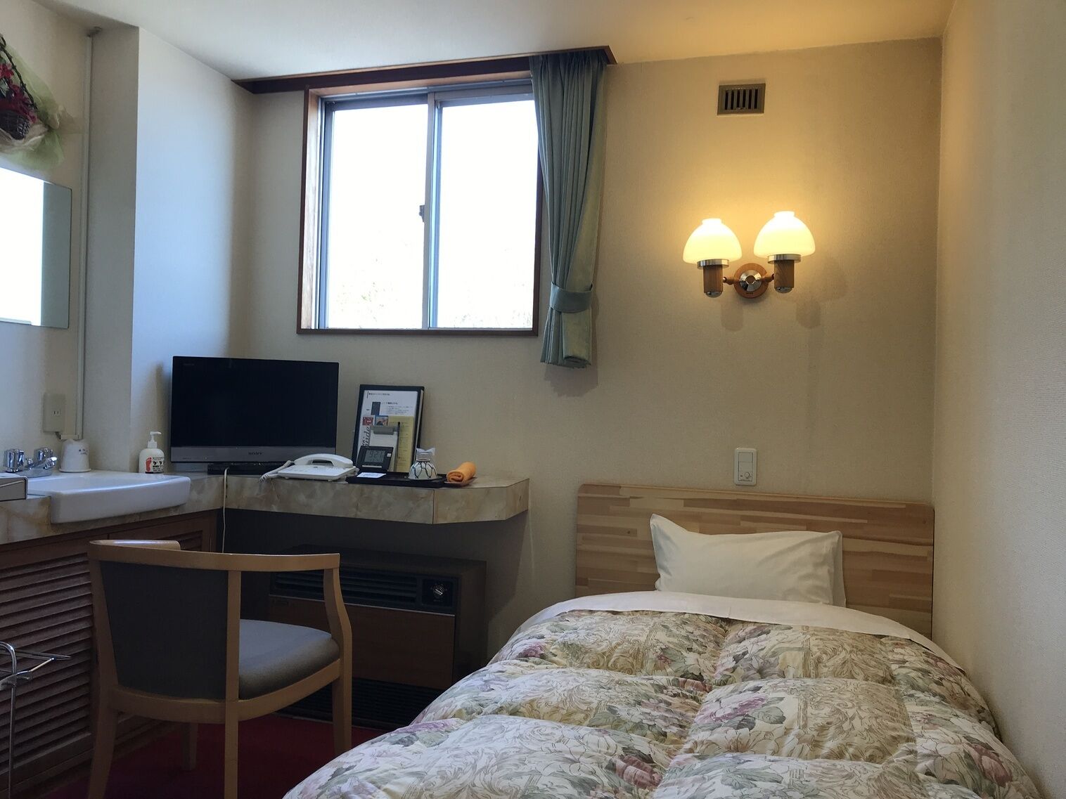 Quarto de hotel com cama, escrivaninha, cadeira e janela com cortinas.