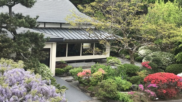 A traditional Japanese-style building with a sloped roof, surrounded by lush greenery and flowering plants. - Hiraizumi Hotel Musashibou (Hiraizumi)