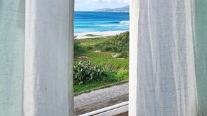 A window with white curtains, revealing a coastal landscape with a beach and ocean under a clear sky. - Terra Nova Cabo Frio Front Beach (Cabo Frio)