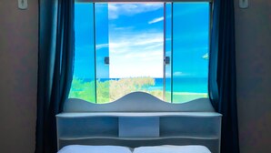 A bed with white pillows and a blue bedspread, a window with a view of the sea and greenery, and dark curtains. - Terra Nova Cabo Frio Front Beach (Cabo Frio)