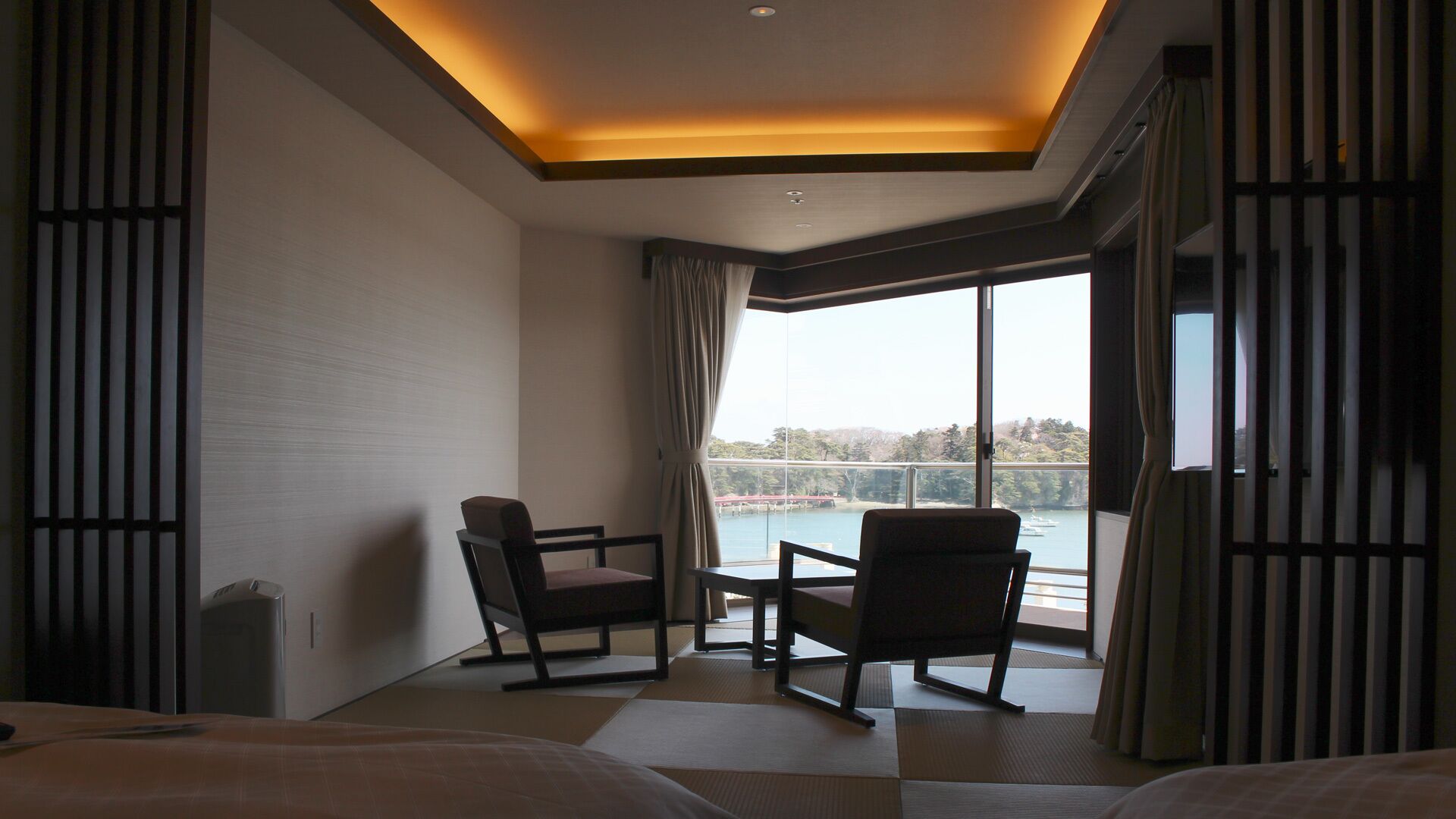 A hotel room with a bed, two chairs, a table, and a large window with a view of a body of water.