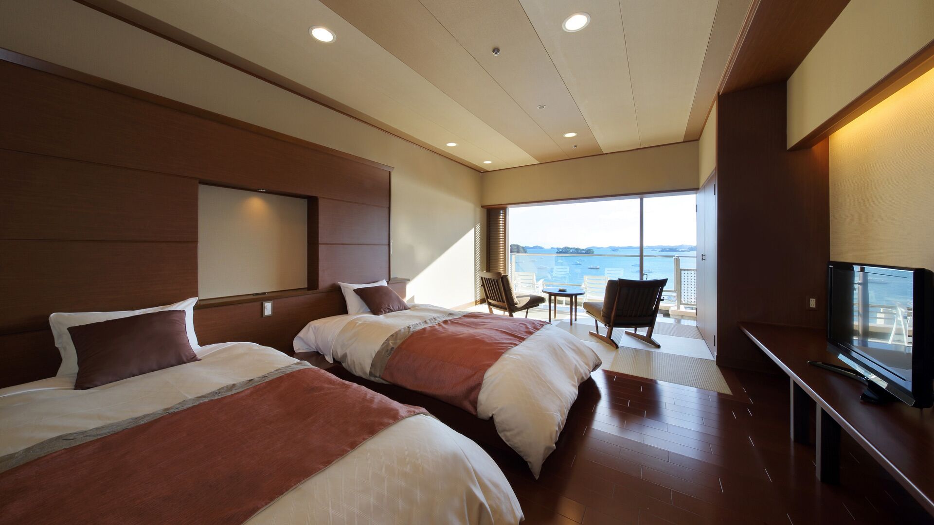 A hotel room with two beds, a TV, a balcony with a view of the sea, and a dining area.