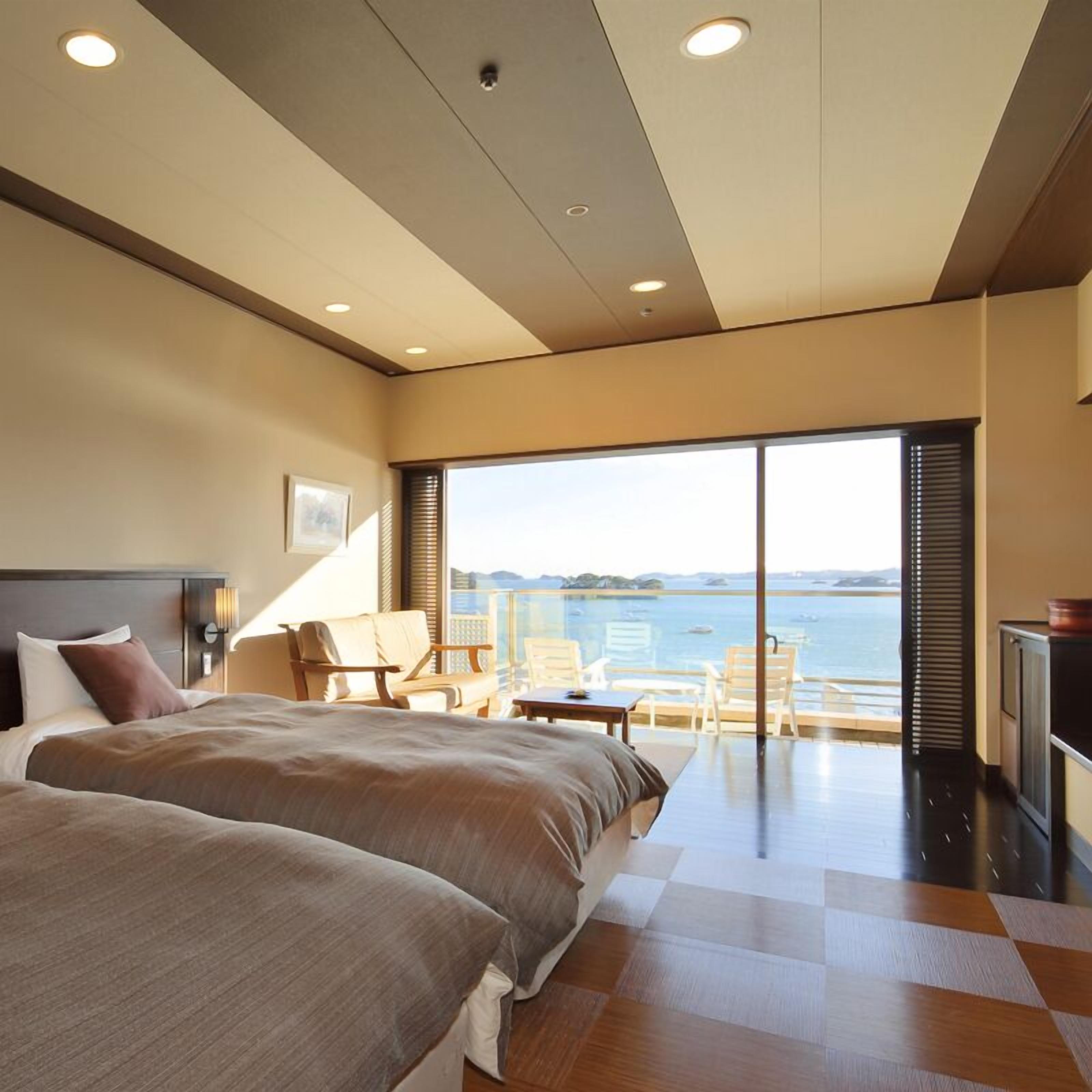 A modern hotel room with two beds, a balcony with a view of the sea, and a large window.