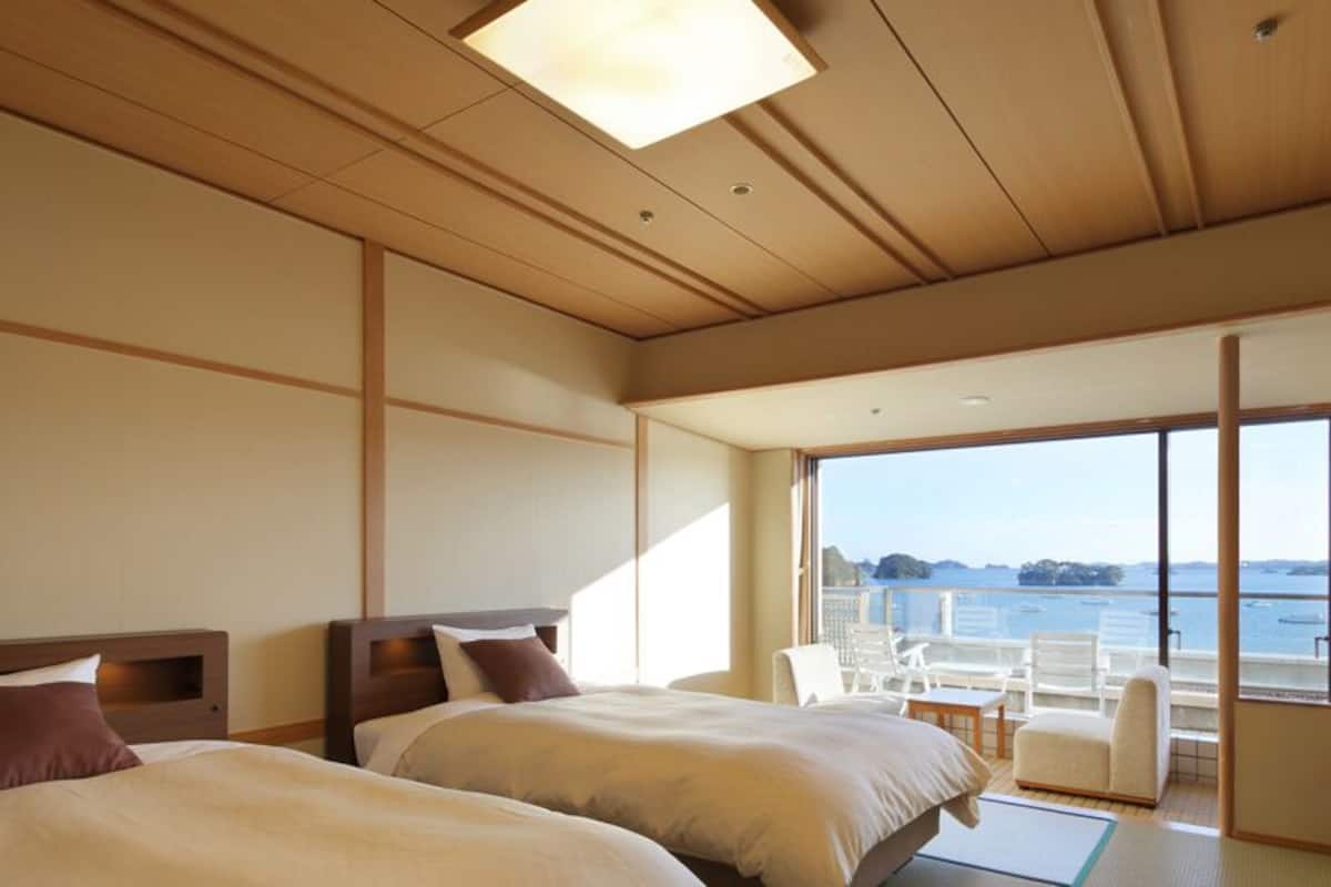 Habitación de hotel con dos camas, un ventanal con vistas al mar y un pequeño rincón de descanso.