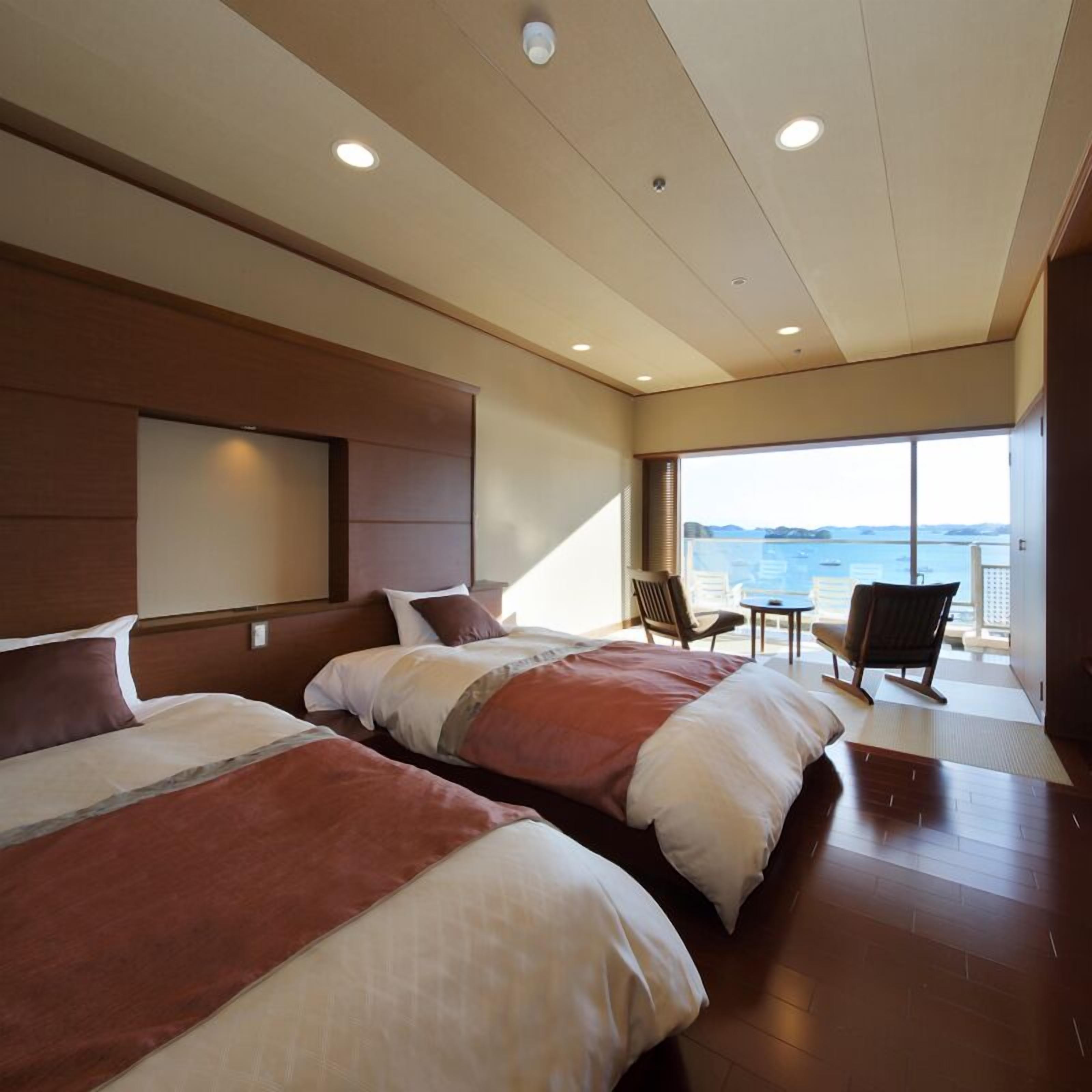A modern hotel room with two beds, a large window with a view of the sea, and a balcony with seating.