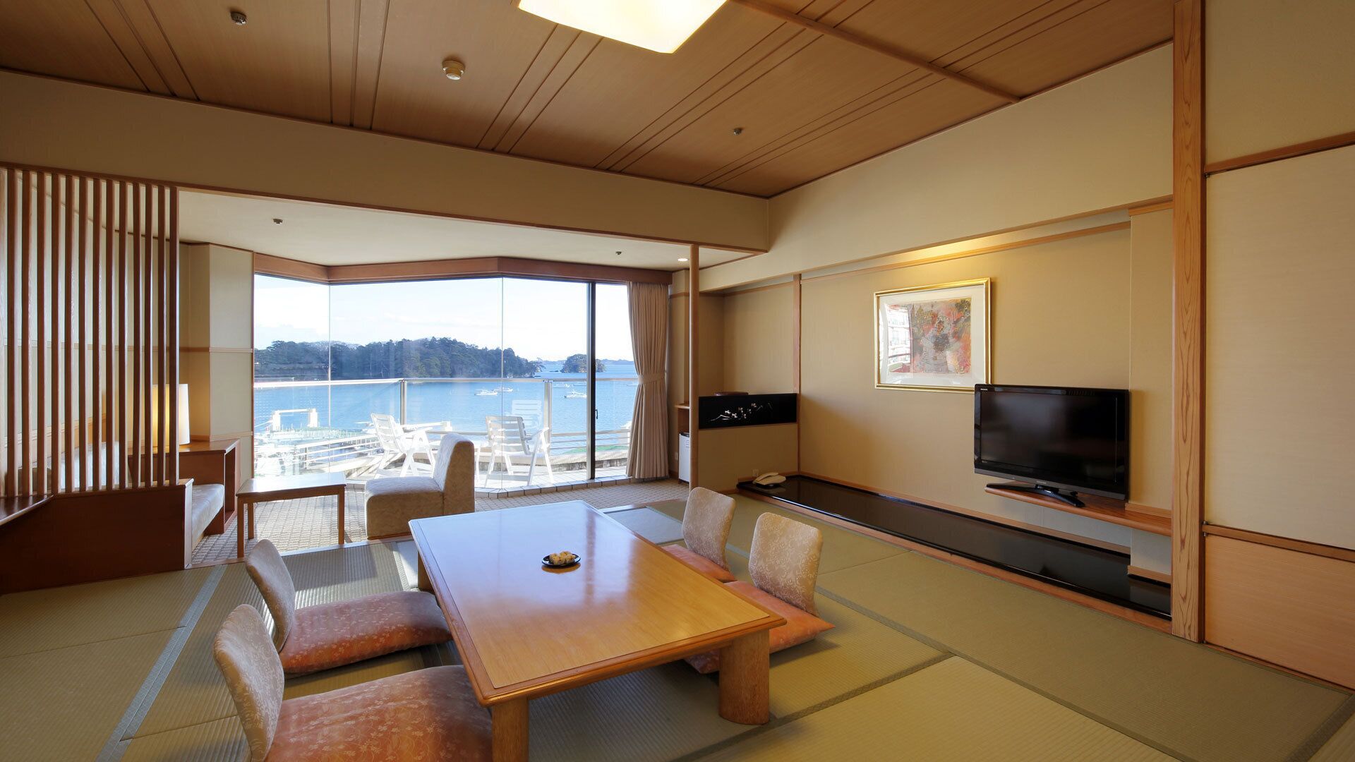 A hotel room with tatami flooring, a low table, cushioned seating, a television, and a large window offering a view of the sea.