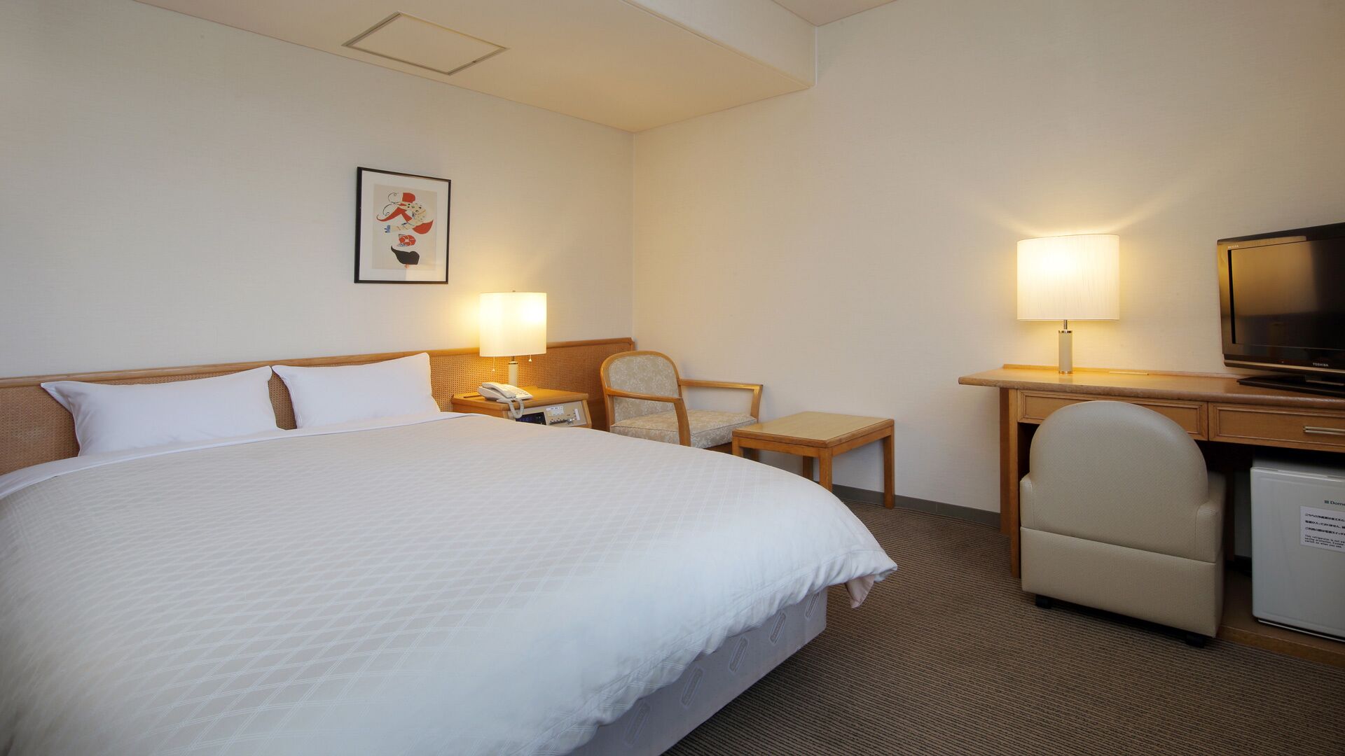 A hotel room with a large bed, a desk with a lamp, a chair, a television, and a small refrigerator.