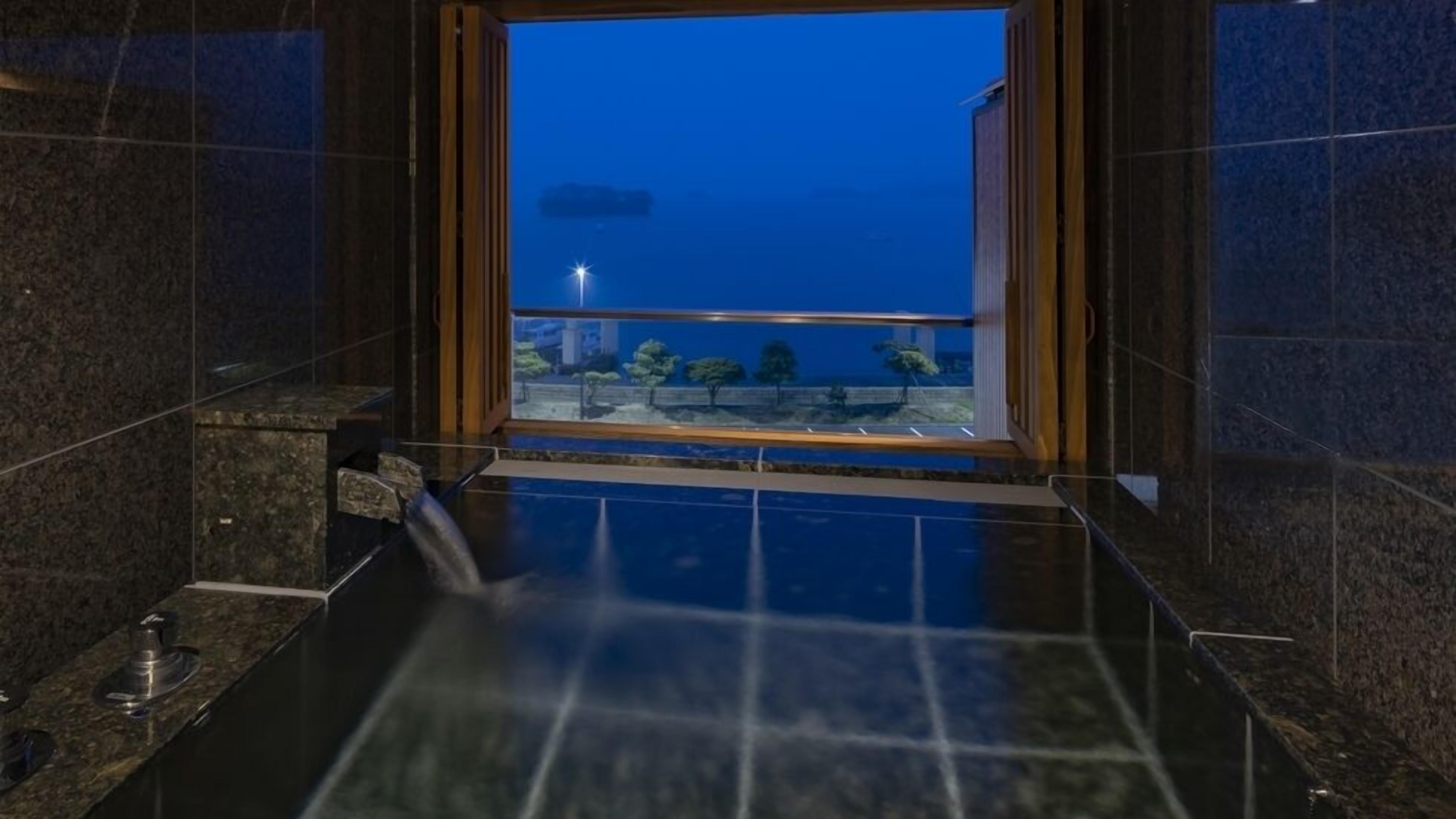A modern bathroom with a large window, a bathtub, and a view of a cityscape at dusk.