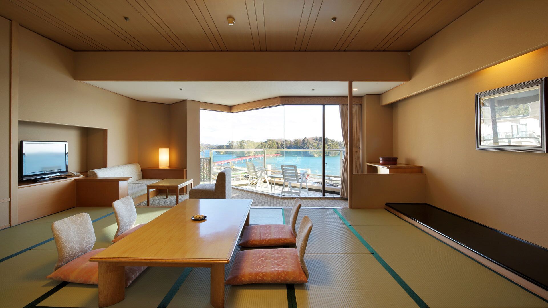 A hotel room with tatami flooring, a dining table, chairs, a TV, a sofa, a balcony with a view of the sea, and a large window.