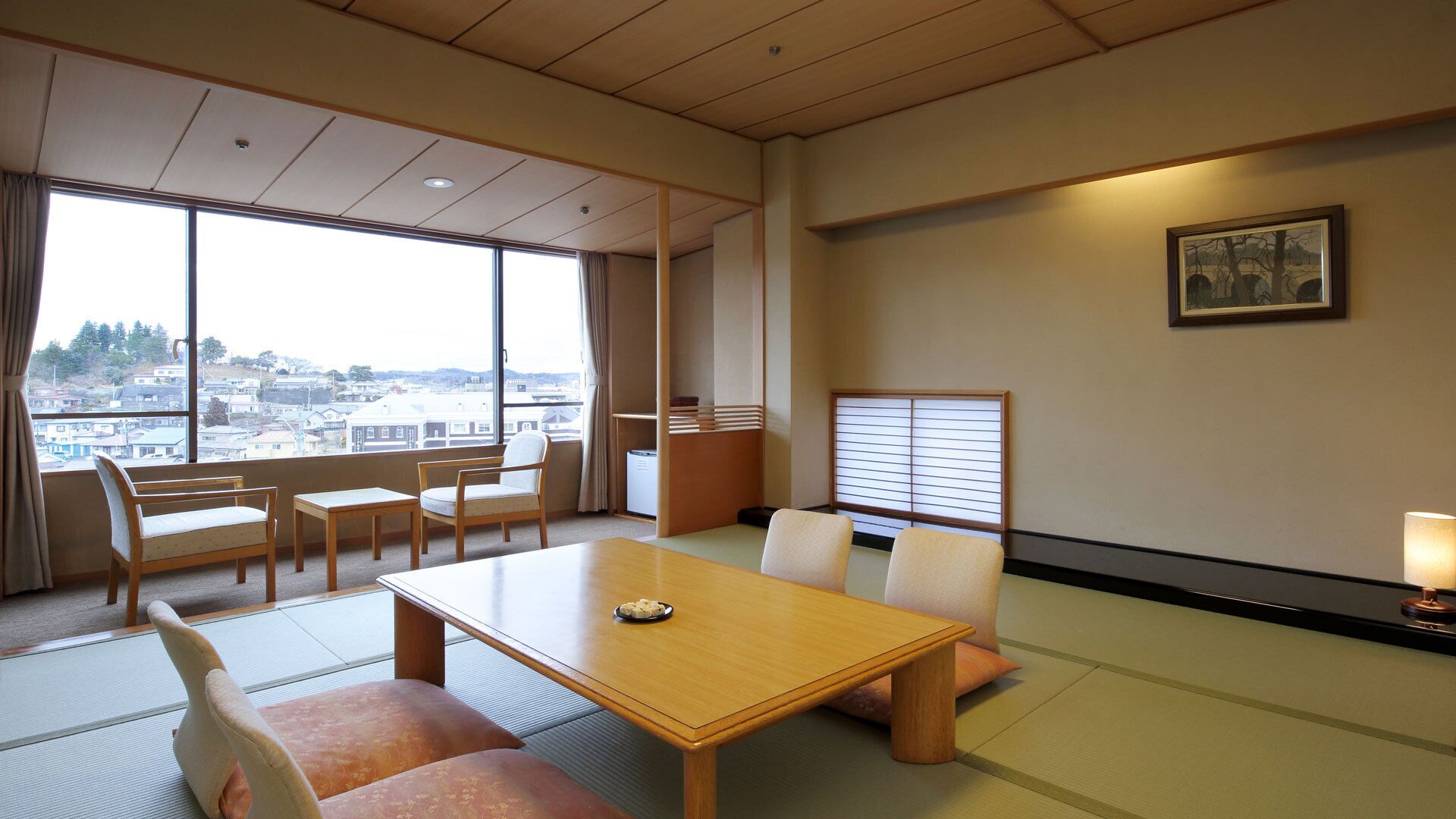 A hotel room with tatami mats, a wooden table, chairs, a view of the city, and a painting on the wall.