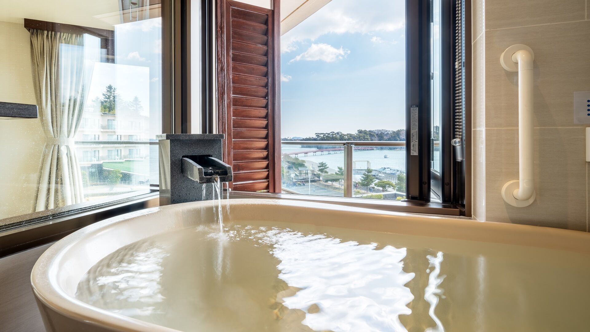 A bathroom with a bathtub, a window with a view of a bridge and water, and a handrail.