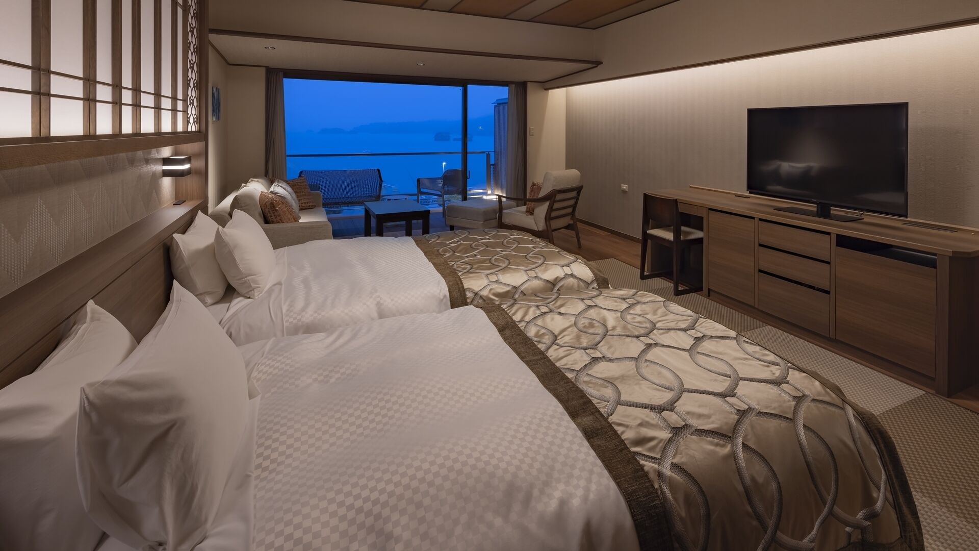 A hotel room with two beds, a TV, a desk, and a balcony with a view.