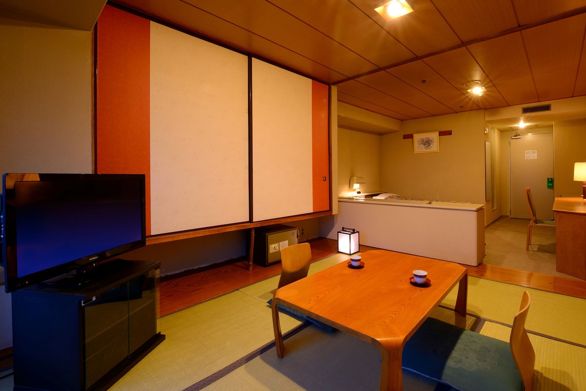 A hotel room with a television, a desk, a chair, a table, and a traditional Japanese floor mat.