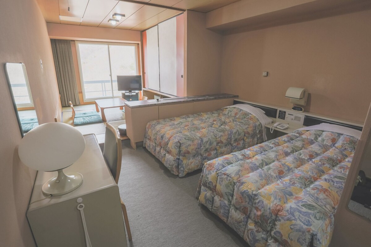 A hotel room with two beds, a desk, a television, and a window.