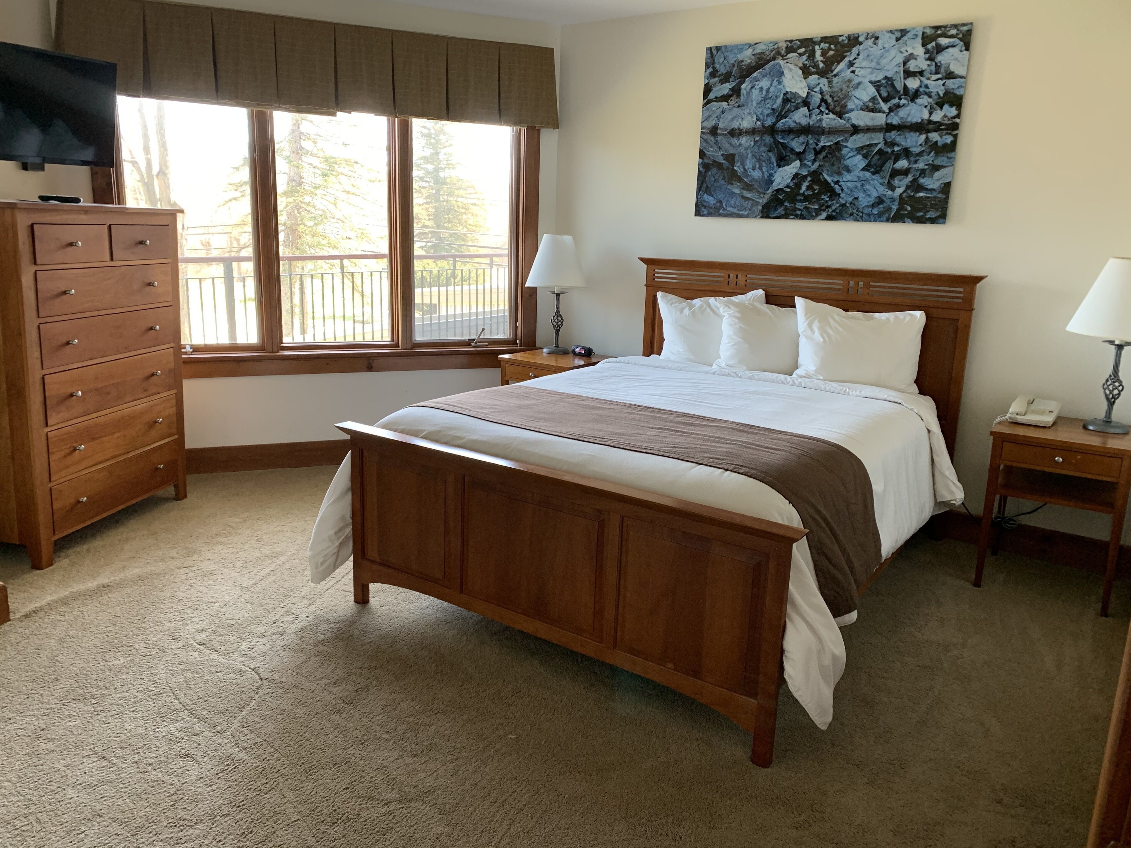 A bedroom with a wooden bed, a dresser, a nightstand with a lamp, a painting on the wall, and a window with blinds.