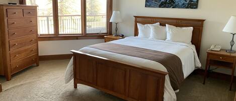 Un dormitorio con cama de madera, cómoda, mesita de noche con lámpara, un cuadro en la pared y una ventana con persianas.
