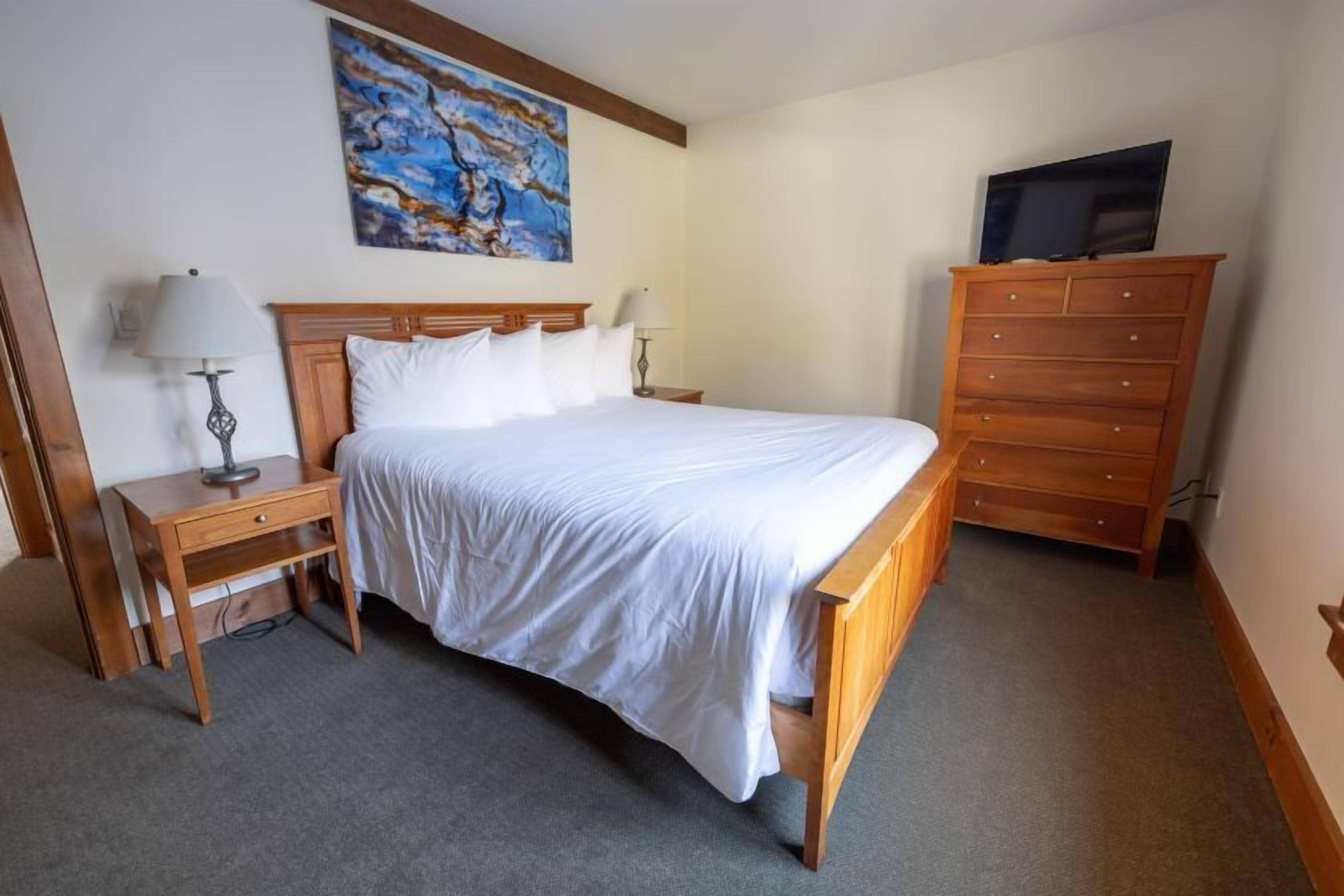 A hotel room with a wooden bed, two bedside tables with lamps, a wooden dresser, and a painting on the wall.