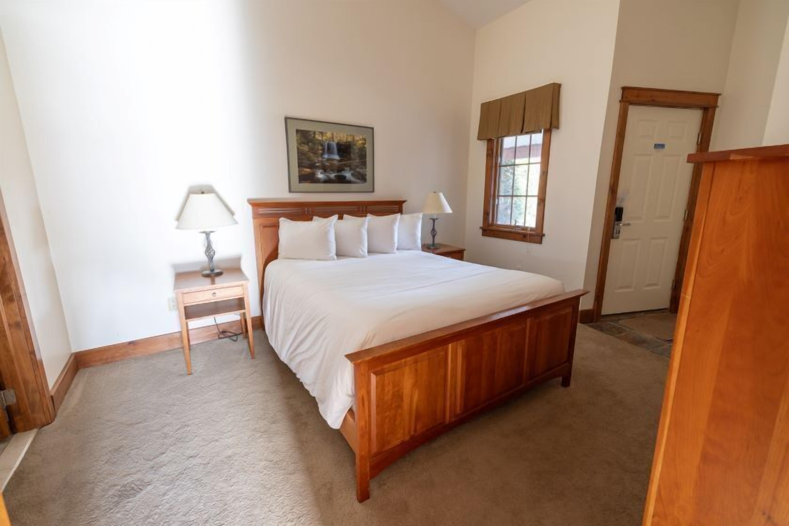 Un dormitorio con una cama de madera, dos mesitas de noche con lámparas, un cuadro en la pared, una ventana con cortinas y una puerta.