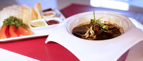 A dining table with a white plate containing a bowl of soup, a small bowl of sauce, and some vegetables.