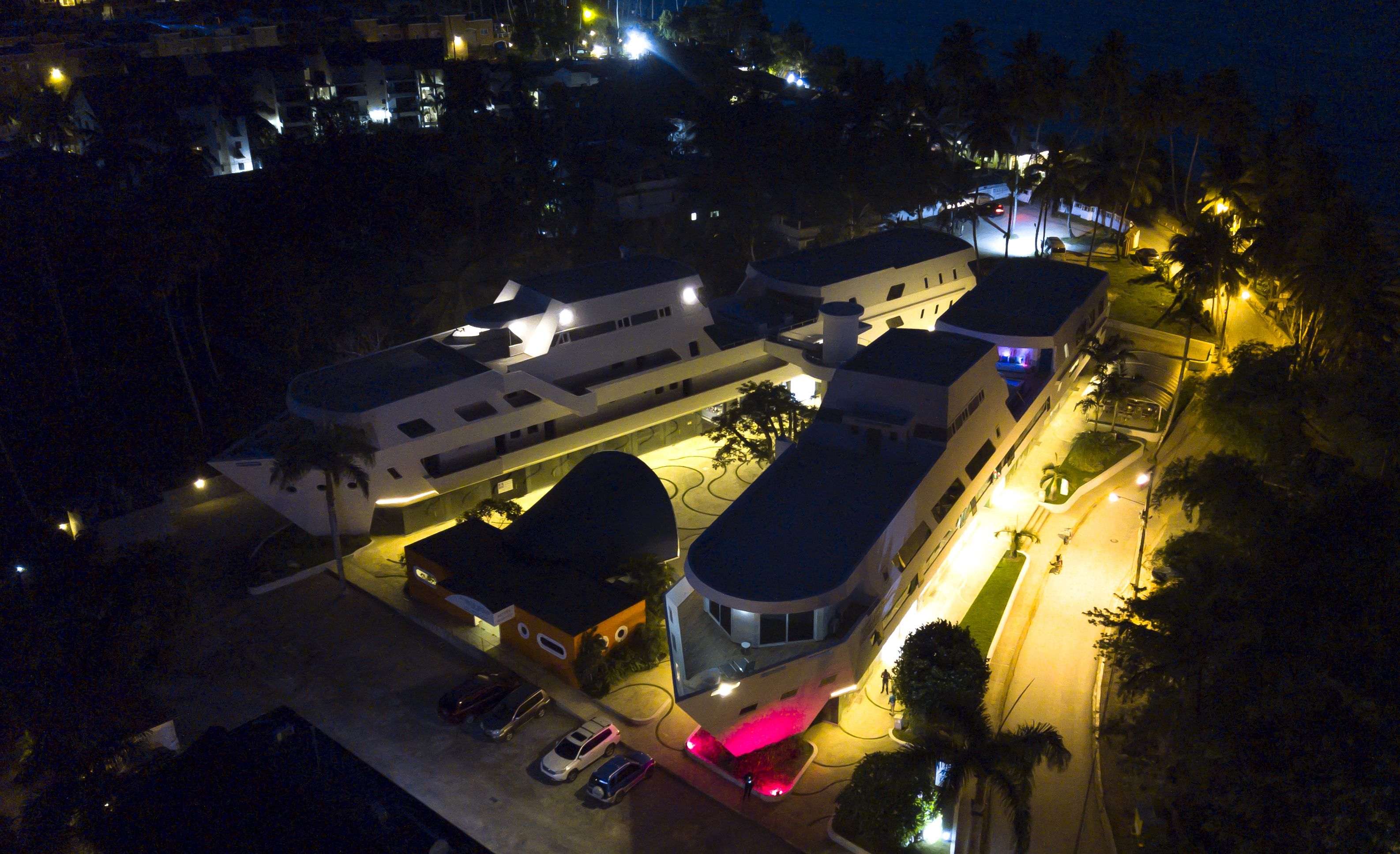 Vista aérea de um moderno conjunto de edifícios, com várias estruturas, incluindo um edifício central imponente com cúpula e alguns edifícios menores. Há estacionamentos com carros e uma rua bem iluminada.
